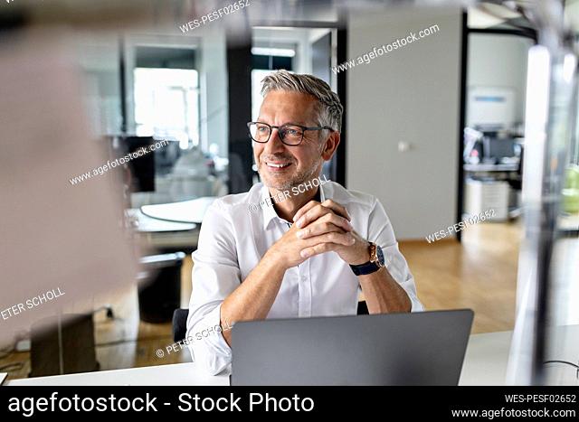 Smiling businessman with hands clasped looking away while sitting at desk in office