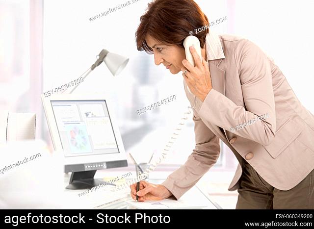 Senior businesswoman at work, standing at desk on landline phone, writing notes