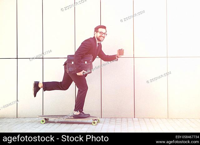 Toned picture of confident businessman smiling for camera and going to his office on longboard. Freelance man holding cup of coffee