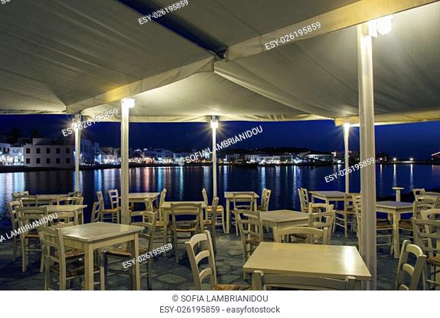 Night view of Chora and the port in Mykonos, Greece. Hora town cityscape lights reflected on the sea, through greek island restaurant with white wooden chairs...