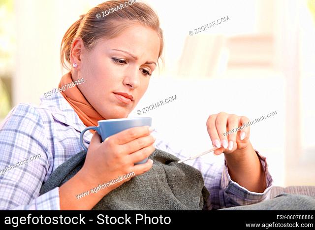 Young woman at home having flu, feeling bad, taking her temperature, drinking hot tea in bed