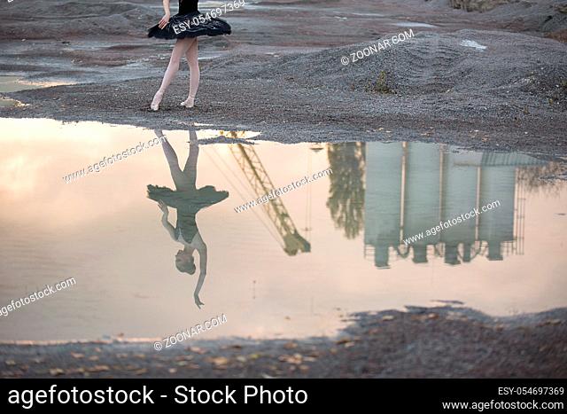 Reflection on the water of a ballerina in a black tutu against the sky, crane and technical structure. She stands on the gravel. Outdoor. Horizontal