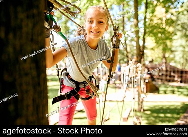 Little smiling girl in equipment climbs in rope park, playground. Child climbing on suspension bridge, extreme sport adventure on vacations
