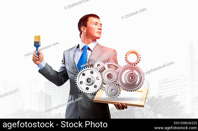 Creative businessman painter holding paint brush and 3d gears mechanism. Portrait of young man in business suit on city background