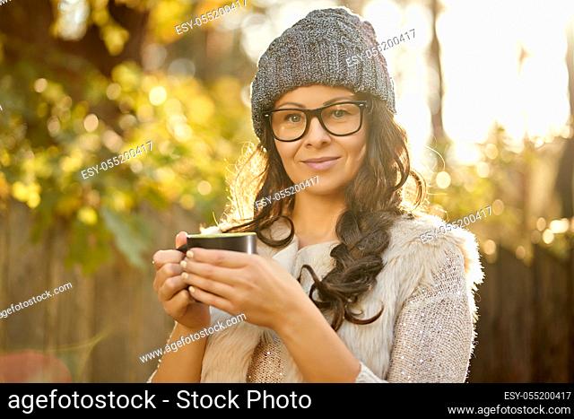 beautiful woman in a cap and glasses holds in her hand a cup of coffee in an autumn forest