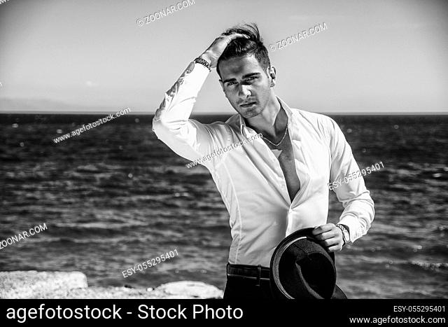 Young handsome man in elegant white shirt and black fedora hat, on beach while looking away. Sea waves on background