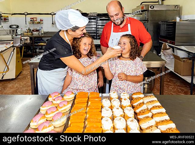 Smiling mother feeding cake to girls while standing with man in bakery kitchen