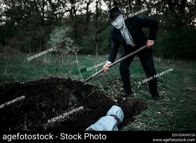 Bloody murderer is digging a grave for the victim in forest, the body wrapped in a canvas, serial maniac concept