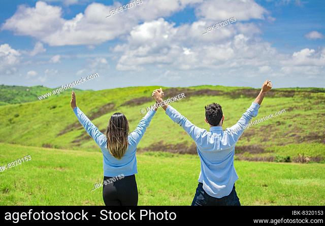 Back view of Happy couple in the field raising hands to the sky, Concept of a happy and free couple in the field, Back view of couple on the hill with hands...