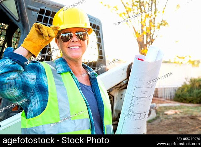 Smiling Female Worker Holding Technical Blueprints Near Small Bulldozer At Constrcution Site