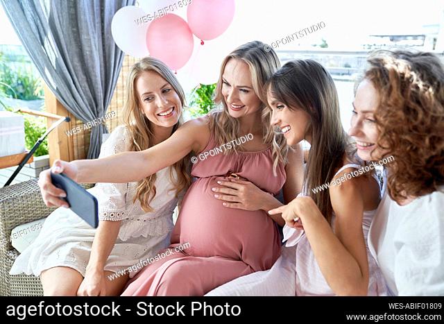 Happy pregnant woman with friends taking selfie through smart phone at baby shower