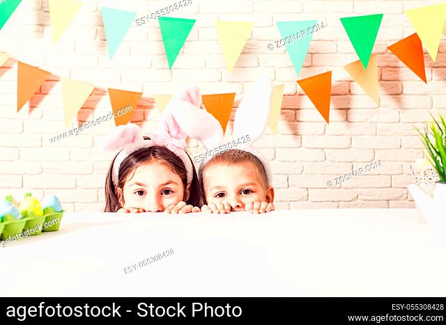 The cute kids with bunny ears peeking from beneath the table. The little boy and girl are waiting for the easter rabbit