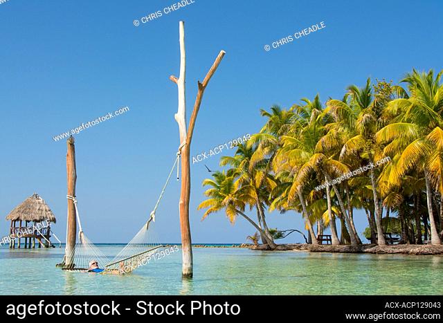 Mature man relaxes in hammock in tropical waters and Palapa platforms, North Long Coco Plum Caye, Belize