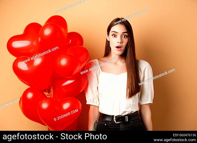 Surprised pretty girl standing near heart balloons and looking with awe at camera. Concept of Valentines day