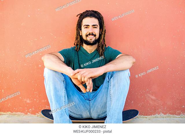 Portrait of smiling young man with dreadlocks and beard sitting on his skateboard in front of a reddish wall
