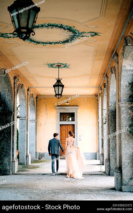 Bride and groom walk along the old terrace with columns entwined with green ivy. Lake Como, Italy. Back view. High quality photo