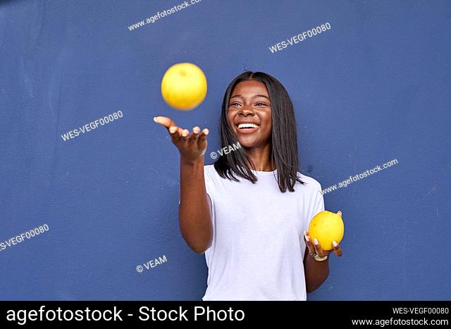 Portrait of happy young woman juggling with two oranges in front of blue background
