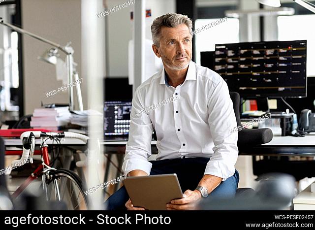 Mature businessman with digital tablet looking away while sitting at office