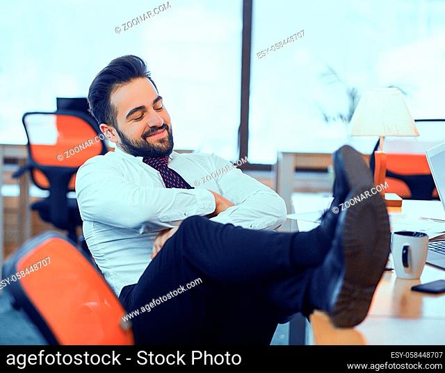 Smiling office worker idle at workplace with his legs thrown on the table. The guy laughs with his arms crossed in front of him