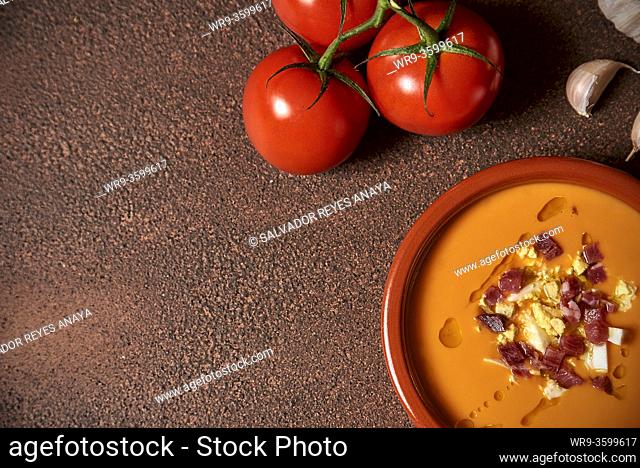 An earthenware bowl with spanish salmorejo, tomatoes, eggs and ham over a rustic wooden table