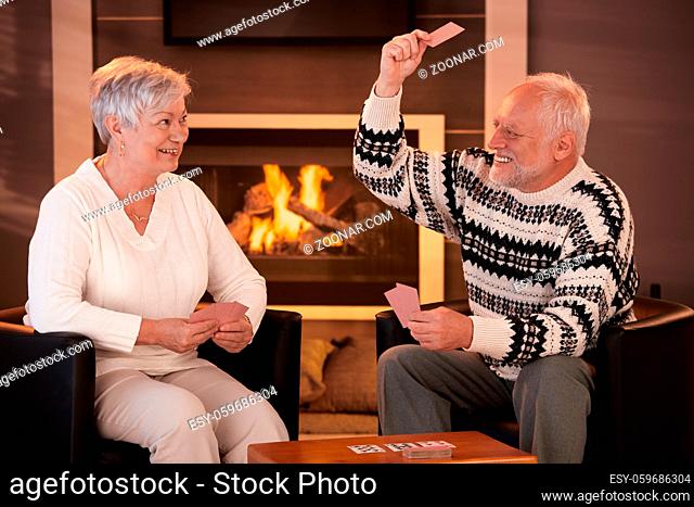 Retired couple playing cards in front of fireplace in living room at home, smiling