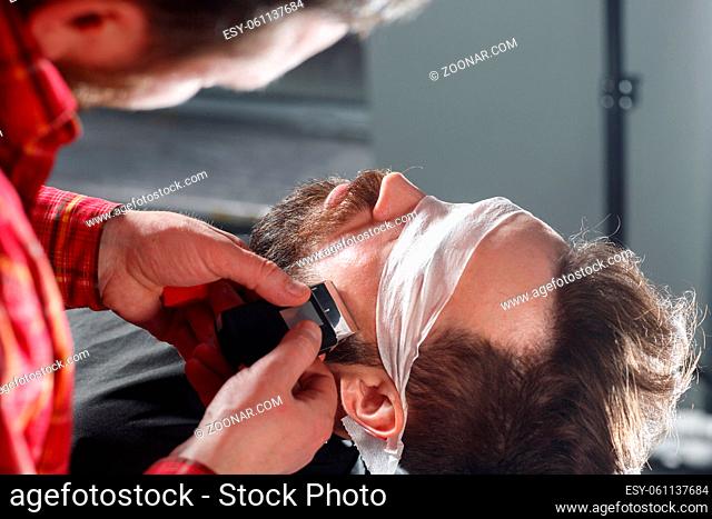 Close-up side view portrait of handsome young bearded caucasian man getting beard grooming in modern barbershop. Making beard haircut using shaver