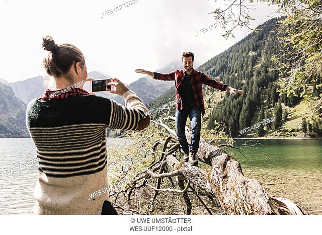Austria, Tyrol, Alps, woman taking cell phone picture of man balancing on tree trunk at mountain lake