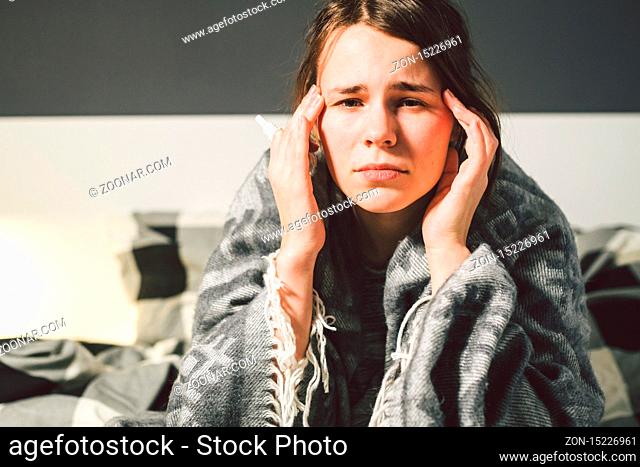 Subject disease and treatment. young Caucasian woman sits home bedroom bed wrapped in body blanket with high fever, fever holds strong headache behind head