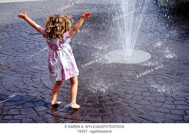 Barefoot little girl with curly hair, in dress, arms wide, enjoying spray from street-level fountain