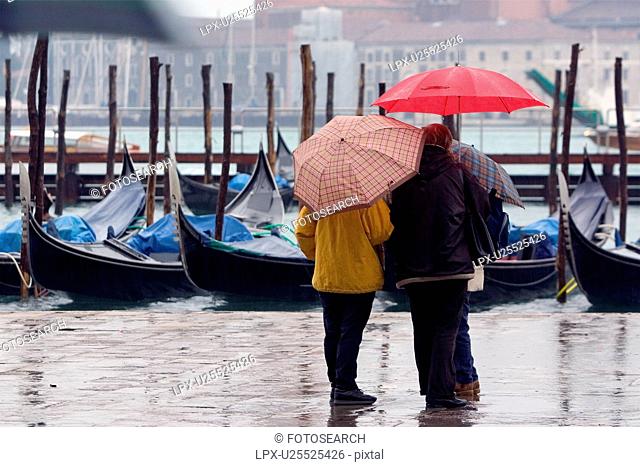 Tourists under umbrellas at edge of St Marks Basin in rain, moored gondola covered with blue tarpaulin in background
