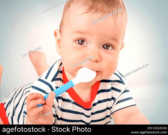 Nine month old baby lying in the bed with spoon. Cute hungry boy in striped bodysuit