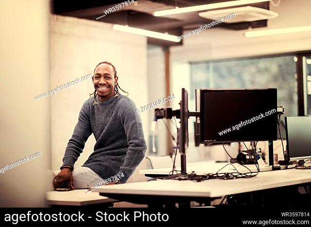 Portrait of young smiling african american male software developer sitting on office desk at modern startup office