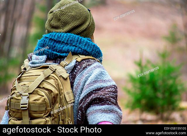 Young woman with layers of heavy, colored winter clothes, blue scarf and green hat from behind, during a walk or a hike in the woodlands