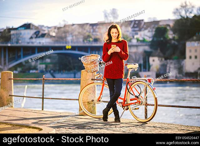 theme tourism on bicycle and modern technology. Beautiful young caucasian woman stands near red retro bicycle on riverside river Rhine Basel Swiss winter warm...