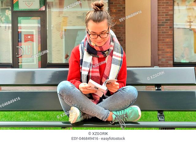 Asian girl in glasses sits on a bench, crosses her legs and reads a book