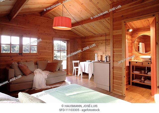 Open-plan living space in a cosy wooden house; a central pendant lamp provides a warm light
