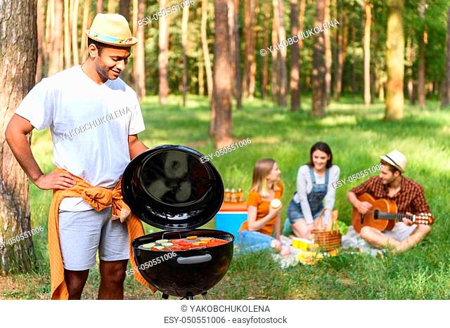 Joyful man is cooking barbecue on grill. He is looking at food with satisfaction and smiling. His friends are sitting and singing to guitar on background