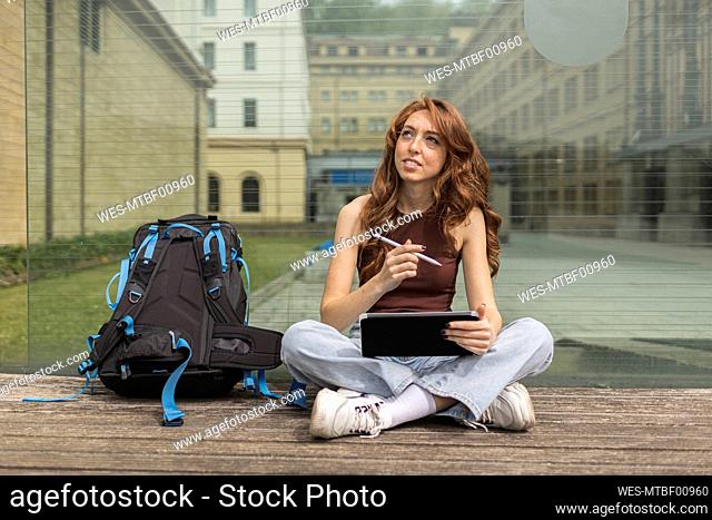 Woman with digital tablet sitting by backpack in front of glass wall