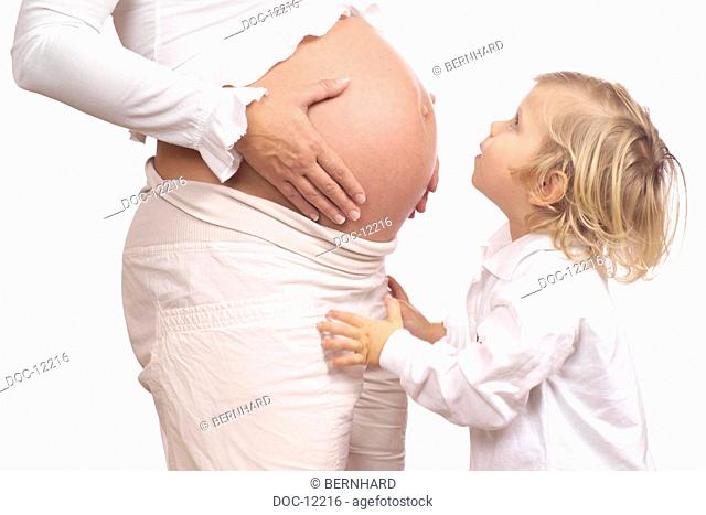 Kid stands before the stomach of a pregnant lady and looks surprisedly highly at it
