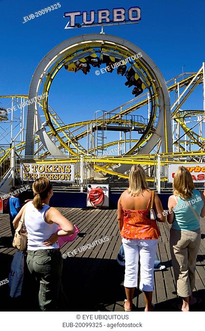 Women watching the Turbo rollercoaster amusement ride on Brighton Pier with the cars full of people upside down at the top of a loop
