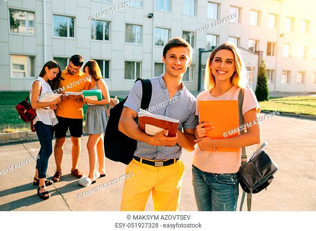 Two happy students with backpacks and books in their hands smiling at camera while standing on background of university and friends