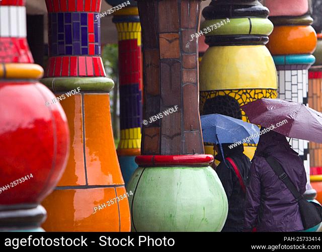 Passers-by protect themselves from the rain with an umbrella on the colorful pillars of the Hundertwasser House ""Green Citadel"" in Magdeburg on Monday (July 4