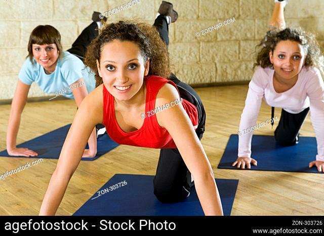 A group of women making stretching exercise on yoga mat. They're smiling and looking at camera. Focus on the woman in red shirt. Front view