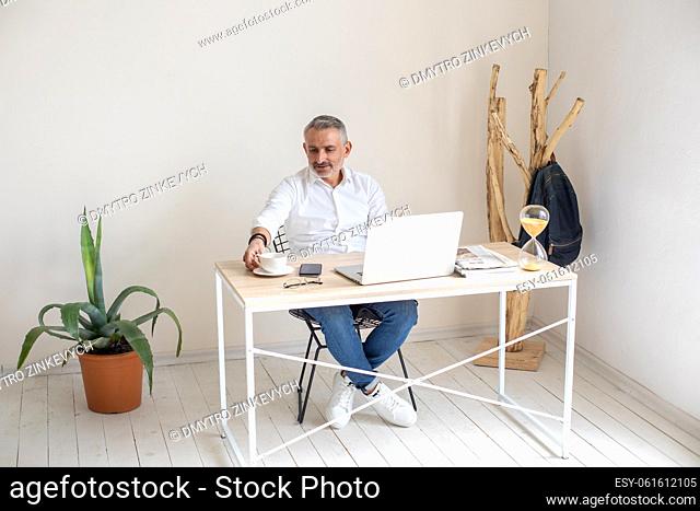 Work, break. Gray-haired stylish man sitting at table with laptop touching cup in modern bright office