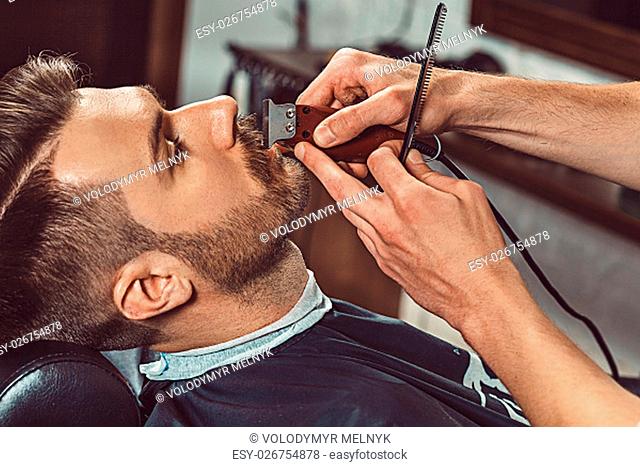 Hipster client visiting barber shop. The hands of young barber making the cut of beard