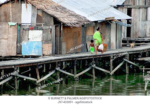 A mother and child drapped in like colored sarongs walk along the common dick of a water village, Sorong Harbour, Western Papua, Indonesia