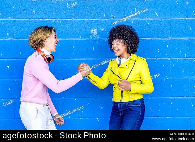 Cheerful multi ethnic friends laughing while holding hand against blue wall