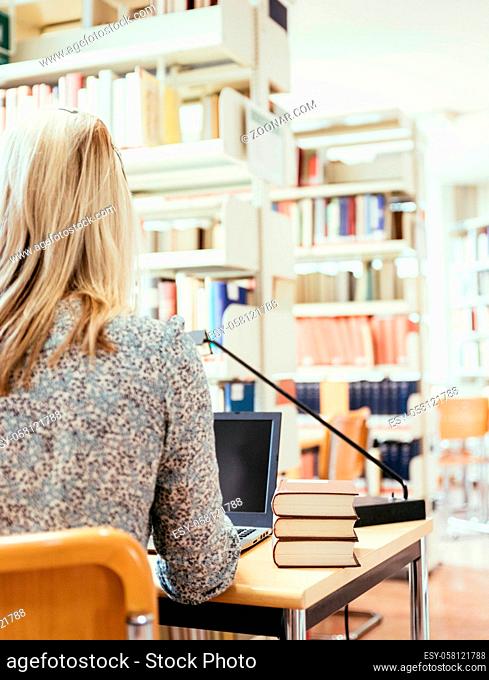Blonde female student is sitting at the desk with pile of books, university library