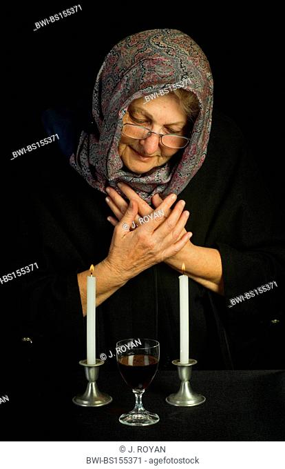An elderly jewish woman, head covered with a scarf, prays over two lit sabbath candles and sabbath wine
