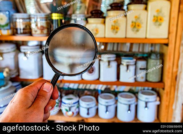 Looking for signs of damp and mold inside the kitchen pantry with a magnifying glass, blurred storage jars are seen displayed on vintage wooden shelves